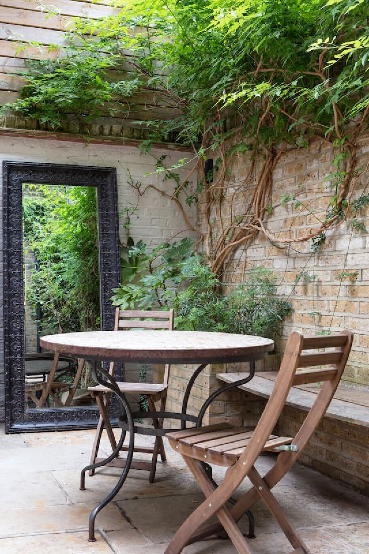 Un espace extérieur aménagé dans un style rustique, avec une table ronde et des chaises en bois, entouré d’un mur de briques et d’une végétation luxuriante.