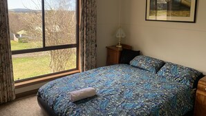 Une chambre avec une grande fenêtre, un lit recouvert d’une couvre-lit à motifs, une table de chevet avec une lampe et un tableau encadré au mur.