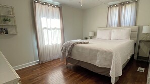 Un dormitorio con cama, ventilador de techo, suelo de madera y ventana con cortinas.
