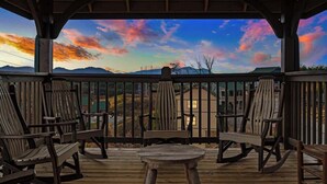 Una terraza de madera con sillas y una mesa, con vistas a un paisaje montañoso y un cielo colorido.