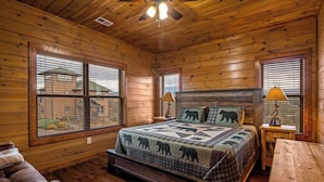 Un dormitorio de cabaña de madera con una cama, dos ventanas, un ventilador de techo y un banco de madera.