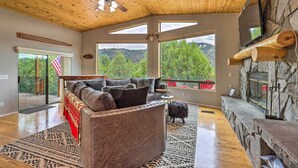 A spacious living room with a stone fireplace, large windows offering a view of the outdoors, a ceiling fan, and a cozy seating area. - 'Elkwood Lodge' Cabin w/ Hot Tub in Star Valley! (Star Valley)