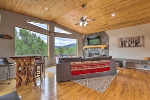 A spacious living area with a wooden ceiling, a stone fireplace, a large window offering a view of trees, and a cozy seating area with a blanket.