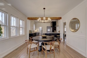 A spacious dining area with a round table and wooden chairs, a chandelier, and large windows.