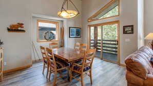 Un espace repas comprenant une table et des chaises en bois, un lustre et de grandes fenêtres donnant sur l’extérieur.
