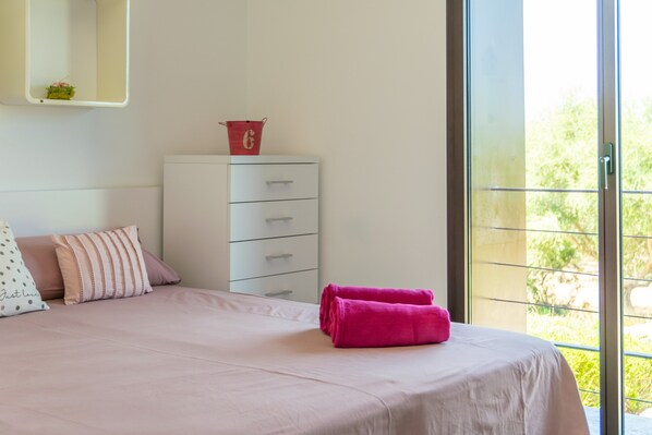A bedroom with a bed, a white dresser, a red bucket, and a view of greenery through a large window. - SA GARRIGA DEN GARAU - Modern villa with wonderful views and private pool. Free WIFI. (Ariany)