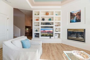 Un salon avec un canapé blanc, un téléviseur à écran plat, une cheminée et une étagère murale ornée d’objets décoratifs.