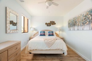 A bedroom with a bed, bedside lamps, a ceiling fan, a dresser, and artwork on the walls.