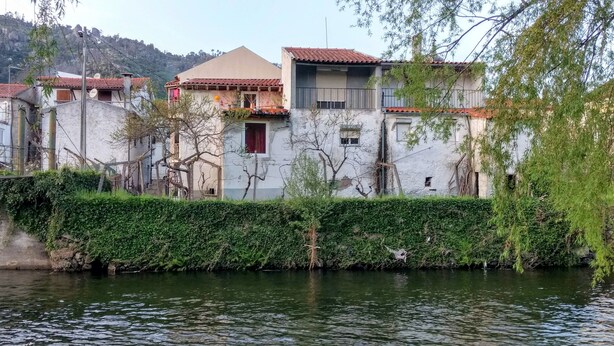 Un edificio junto al río, con fachada blanca, techos de tejas rojas y un seto delantero.
