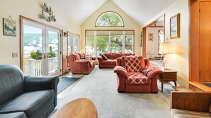 A spacious living room with a high ceiling, a large arched window, and a view of the outdoors. There are multiple seating areas with sofas and a coffee table. - Loon Landing - Fisherman's Paradise on West Shore of Keuka Lake (Hammondsport)