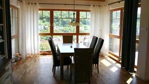 Un coin repas avec une table et des chaises en bois, de grandes fenêtres avec des rideaux et une vue sur la verdure à l’extérieur.