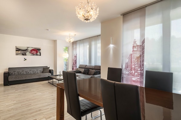 Una sala de estar moderna con un sofá, una mesa de comedor con sillas, una araña y un ventanal con cortinas.