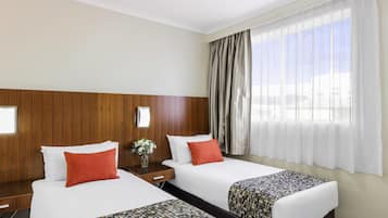 Una habitación de hotel con dos camas, un cabecero de madera, una mesita de noche con un florero y un ventanal con cortinas translúcidas.