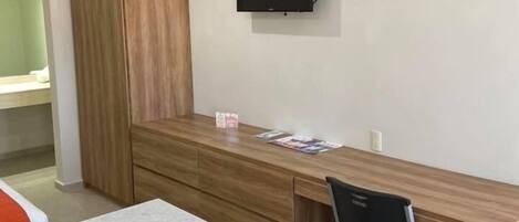 Una habitación moderna y bien iluminada con un escritorio de madera, una silla negra, televisión integrada y una pequeña área de kitchenette con lavavajillas.