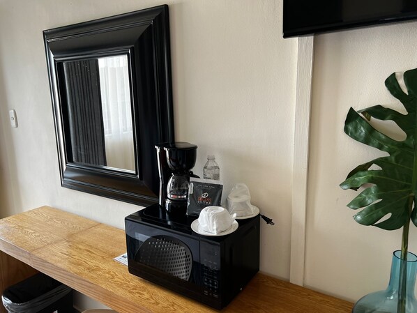 A coffee maker, a water bottle, a disposable cup, and a plant on a wooden table in a room with a mirror and a television. - Hotel Los Robles (Nuevo Laredo)