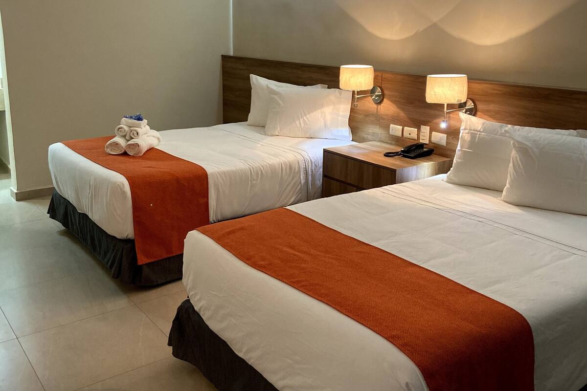 A hotel room with two beds, each with white linens and orange throw blankets. There are two bedside tables with lamps and a phone.