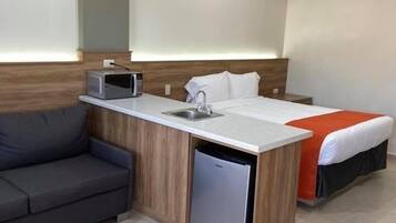 A compact hotel room with a kitchenette, a bed with an orange accent, a grey sofa, and a small refrigerator.