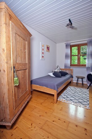 A bedroom with a wooden wardrobe, a bed with a grey mattress, a small round table, and a window with curtains.