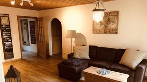 En hyggelig stue med en mørkebrun lædersofa, et træbord og et stort gulvtæppe. Der er et kort på væggen og en trædør, der fører til et andet rum.