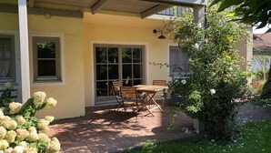 En overdækket terrasse med et træbord og stole, en plantekasse og et vindue med udsigt til haven.