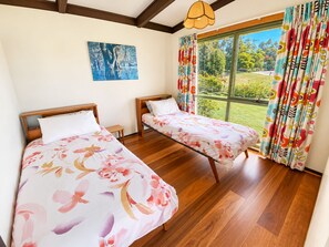 Une chambre avec deux lits, un plancher en bois, une grande fenêtre avec des rideaux et un tableau au mur.