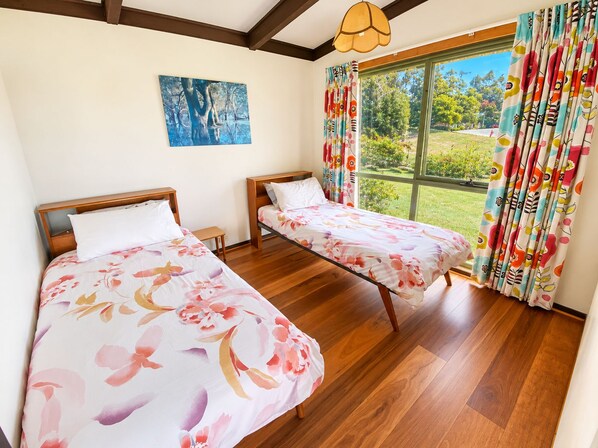 A room with two beds, wooden flooring, a large window with curtains, and a painting on the wall. - Sylvin Waters, Mallacoota (Mallacoota)