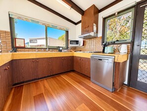 A modern kitchen with wooden cabinets, a dishwasher, and a window overlooking a residential area. - Sylvin Waters, Mallacoota (Mallacoota)