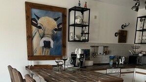 Een keuken met een houten kookeiland, barstoelen, een schilderij van een koe en een zwarte plankenkast met servies.