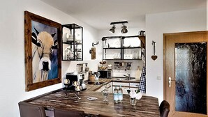 Een eetgedeelte met een houten tafel en stoelen, een keuken op de achtergrond, en een schilderij aan de muur.