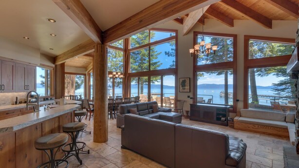 A spacious living area with a large window overlooking a lake, wooden beams on the ceiling, and a bar area with stools.