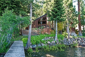 A cabin with a wooden deck leading to a stone wall and a small pond. - Cathedral Lakefront by Tahoe Getaways - 6 BR Estate w Pier, Buoy and Pool Table (Tahoe City)