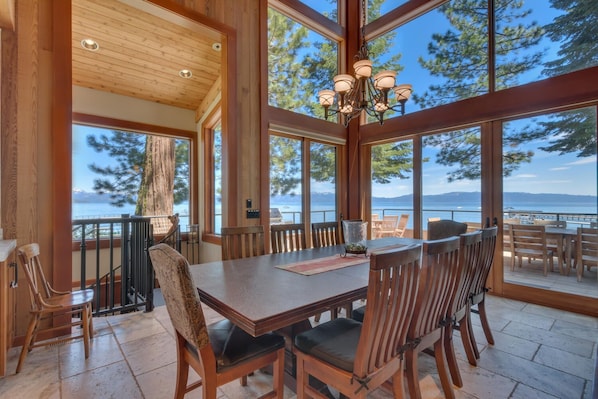 A wooden dining area with a large table and chairs, large windows with a view of the sea and mountains, and a chandelier. - Cathedral Lakefront by Tahoe Getaways - 6 BR Estate w Pier, Buoy and Pool Table (Tahoe City)