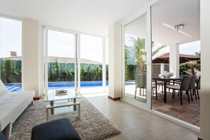 A modern living room with a glass door leading to a pool area, a white sofa, a glass coffee table, and a dining area with outdoor furniture.