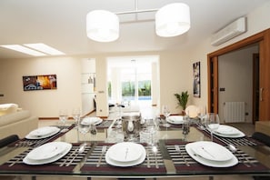 A dining area with a glass table set for four, surrounded by chairs. There are two pendant lights above the table, and a large window in the background.