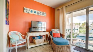 Een kamer met een televisie, rieten meubels, een rieten stoel, een rieten bank, een rieten tafel, een rieten mand en uitzicht op een zwembad en groen buiten.