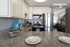 A kitchen with white cabinets, a stainless steel refrigerator, a microwave, and a toaster. There are two wine glasses on the granite countertop. - On the Beach... No need to cross the Busy Seawall (Galveston)