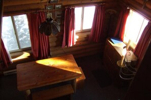 Ein Interieur einer Holzhütte mit einem Holztisch, einer Hängelampe und einem Fenster mit Vorhängen.