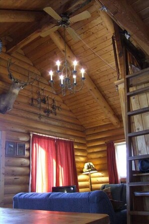 A log cabin interior with a chandelier, red curtains, a ceiling fan, and a ladder.
