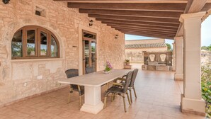 An outdoor patio with a stone wall, wooden beams, a table with chairs, and a view of a traditional building in the background. - Sa Punta Ses Salines Holiday Home with Pool and Wi-Fi (Ses Salines)