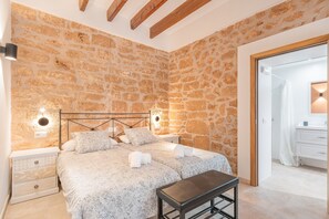 A bedroom with a stone wall, a bed with patterned bedding, bedside lamps, a bench, and a wooden ceiling.