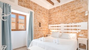 A bedroom with a stone wall, a bed with white bedding, a bench, and a nightstand. - Sa Punta Ses Salines Holiday Home with Pool and Wi-Fi (Ses Salines)