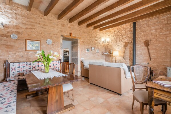 A rustic interior with exposed stone walls, wooden beams, a dining table with chairs, a sofa, and a fireplace.
