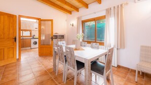 Uma área de refeições com uma mesa e cadeiras brancas, uma janela com cortinas e uma cozinha ao fundo.
