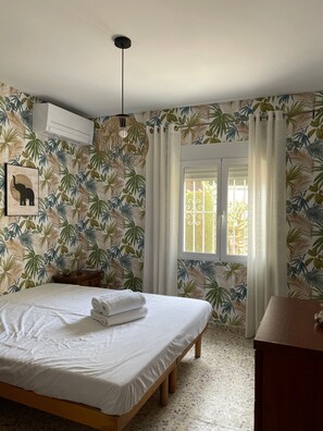 A bedroom with a bed, a bedside table, a wall-mounted air conditioner, a window with curtains, and a tropical leaf-pattern wallpaper. - Villa Casa dos Leones with sea view, pool, air conditioning, Wi-Fi, garden & terrace (Benitachell)