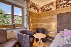 A room with a large window, two plush armchairs, a small wooden table, a bed with floral bedding, and a wall with a wooden panel design.