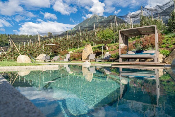 A clear pool with a wooden deck, lounge chairs, and a rock garden. - Apartment "Feldhof Apfeltraum" with Saltwater Pool, Panoramic Lawn and Balcony (Kuens bei Meran)