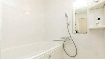 A modern bathroom with a white bathtub, a handheld showerhead, and a mirror above a shelf with toiletries.