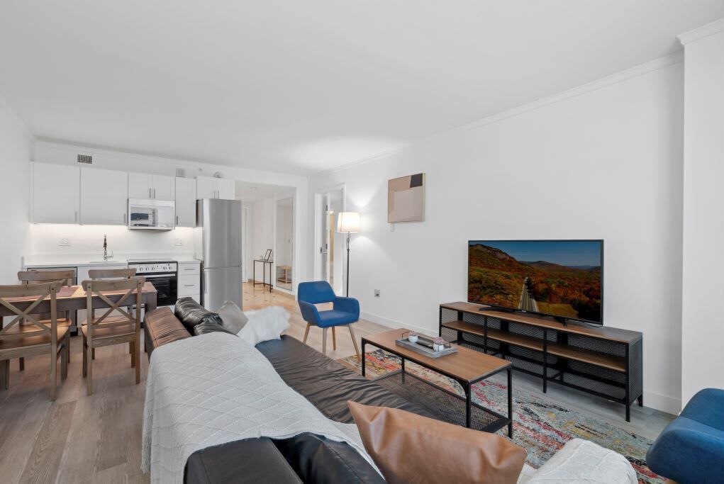 A modern, well-lit living room with a sofa, a coffee table, and a TV. There is a dining area with a table and chairs, and a kitchen in the background.