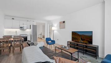 A modern, well-lit living room with a sofa, a coffee table, and a TV. There is a dining area with a table and chairs, and a kitchen in the background.