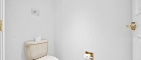 A small, white bathroom with a toilet, a gold flush handle, and a white toilet paper holder.